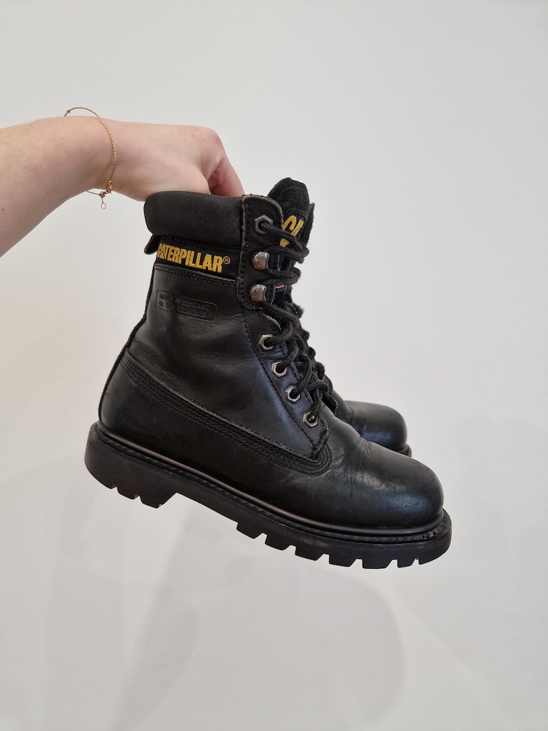 Puede incluir: Botas de trabajo de cuero negro con el nombre de la marca "Caterpillar" en letras amarillas. Las botas tienen un dise&ntilde;o con cordones, un cuello acolchado y una suela gruesa y resistente. Las botas est&aacute;n sujetas por una mano.