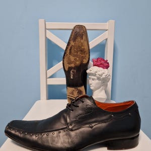 Puede incluir: Zapato de vestir de cuero negro con puntera cuadrada y detalles con tachuelas. El zapato tiene cierre de cordones y un dise&ntilde;o decorativo en el lateral. La suela presenta una ilustraci&oacute;n detallada. Se ve un segundo zapato, suela hacia arriba.