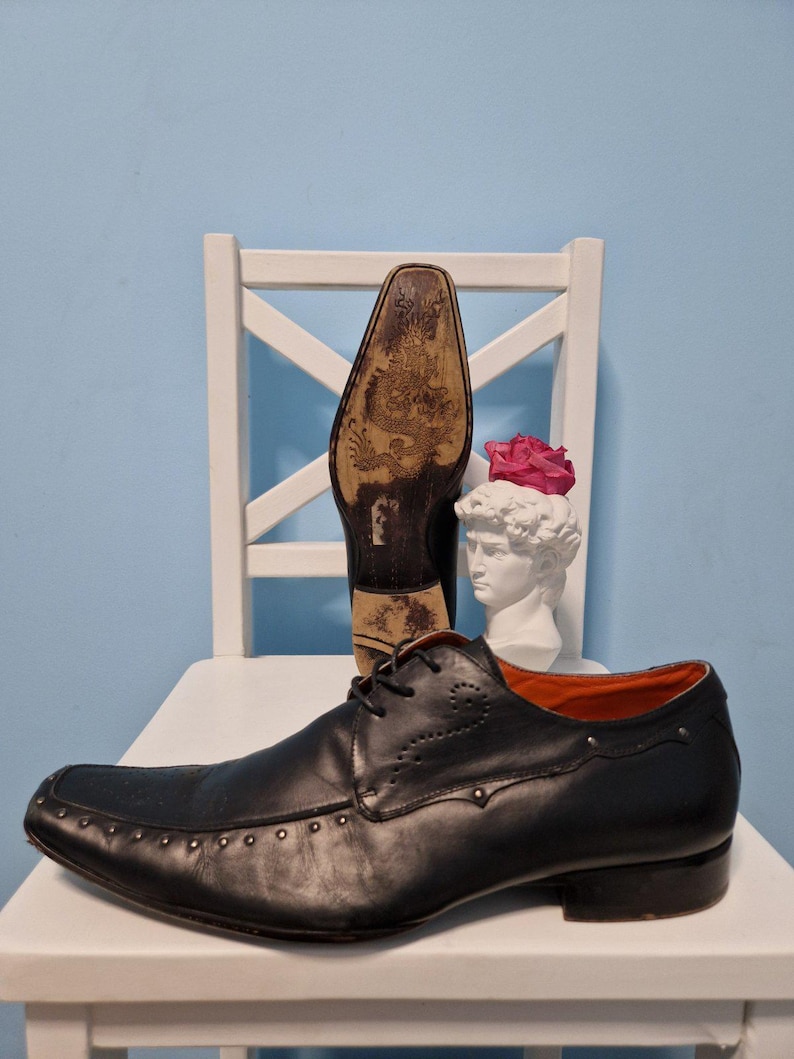 Puede incluir: Zapato de vestir de cuero negro con puntera cuadrada y detalles con tachuelas. El zapato tiene cierre de cordones y un dise&ntilde;o decorativo en el lateral. La suela presenta una ilustraci&oacute;n detallada. Se ve un segundo zapato, suela hacia arriba.