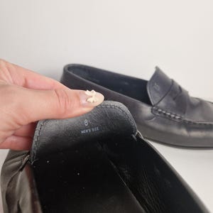 May include: Black leather loafers with a classic design. The shoe's interior shows the size 8 and the words "MEN'S SIZE". The second shoe is visible in the background. The shoes are on a white background.