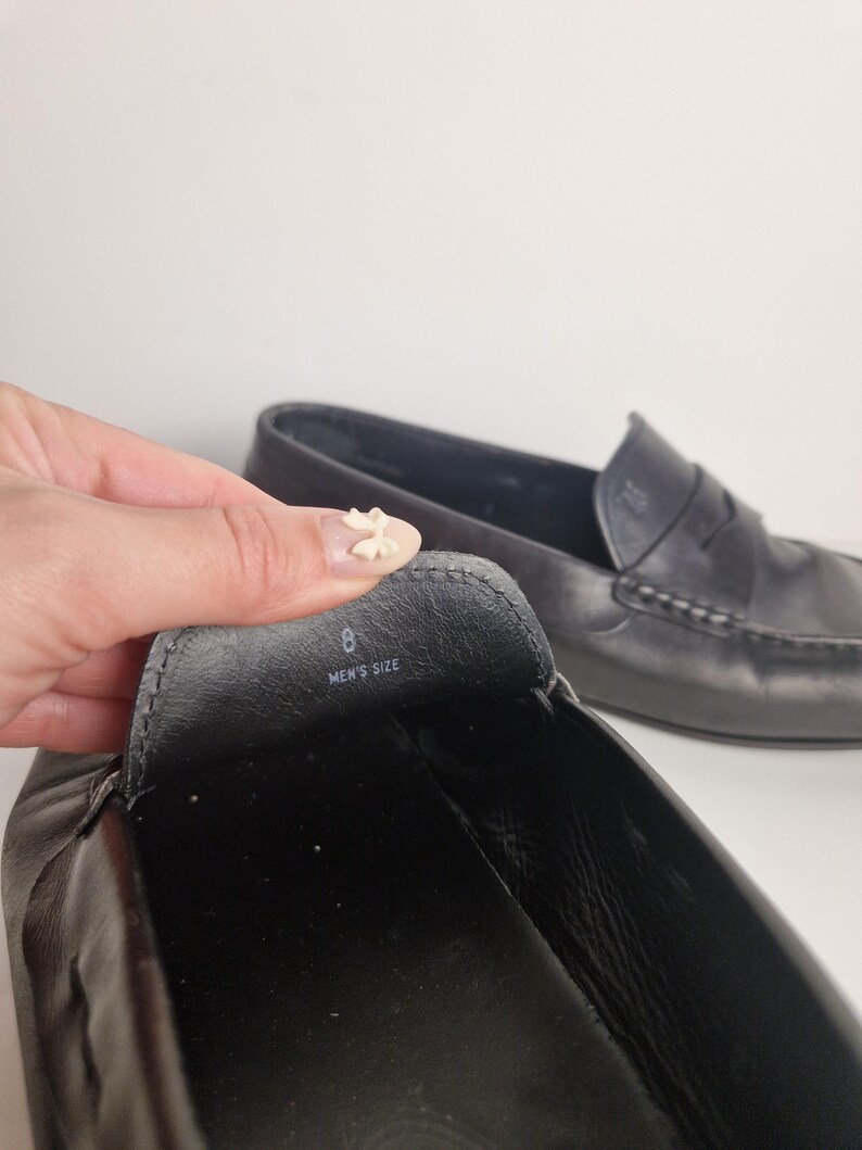 May include: Black leather loafers with a classic design. The shoe's interior shows the size 8 and the words "MEN'S SIZE". The second shoe is visible in the background. The shoes are on a white background.