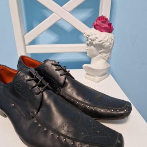 Puede incluir: Un par de zapatos de vestir de cuero negro con tachuelas plateadas y punta cuadrada. Los zapatos tienen cierre de cordones y se exhiben sobre una superficie blanca. Una escultura de busto blanco con una rosa rosa en el fondo.