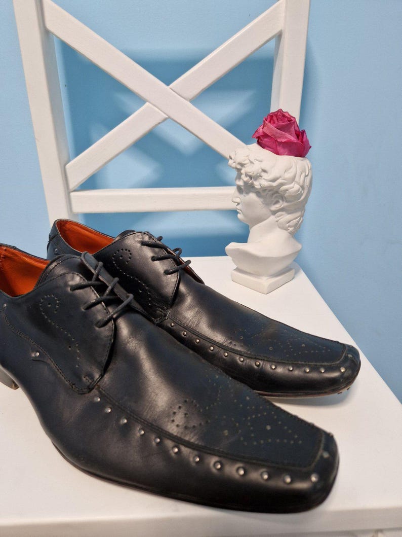 Puede incluir: Un par de zapatos de vestir de cuero negro con tachuelas plateadas y punta cuadrada. Los zapatos tienen cierre de cordones y se exhiben sobre una superficie blanca. Una escultura de busto blanco con una rosa rosa en el fondo.