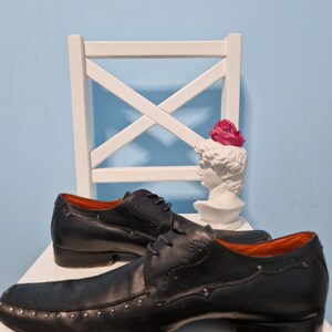 Puede incluir: Un par de zapatos de vestir de cuero negro con tachuelas decorativas en la puntera y los laterales. Los zapatos tienen un dise&ntilde;o cl&aacute;sico con cordones y un tac&oacute;n bajo. El interior es naranja. Un busto blanco con una flor rosa est&aacute; en el fondo.