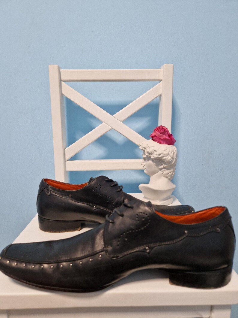 Puede incluir: Un par de zapatos de vestir de cuero negro con tachuelas decorativas en la puntera y los laterales. Los zapatos tienen un dise&ntilde;o cl&aacute;sico con cordones y un tac&oacute;n bajo. El interior es naranja. Un busto blanco con una flor rosa est&aacute; en el fondo.