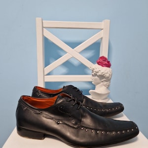 Puede incluir: Un par de zapatos de vestir de cuero negro con tachuelas plateadas en la puntera y los laterales. Los zapatos tienen un dise&ntilde;o cl&aacute;sico con cordones y est&aacute;n colocados sobre una silla blanca sobre un fondo azul claro.