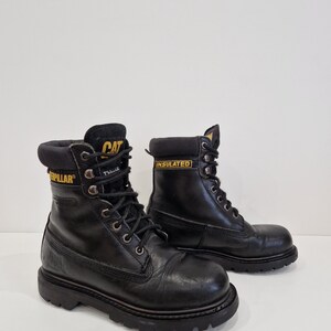 Puede incluir: Botas de trabajo de cuero negro con letras amarillas "CAT" e "INSULATED". Las botas tienen un dise&ntilde;o con cordones, ojales met&aacute;licos y una suela gruesa. Botas dise&ntilde;adas para el trabajo y actividades al aire libre.