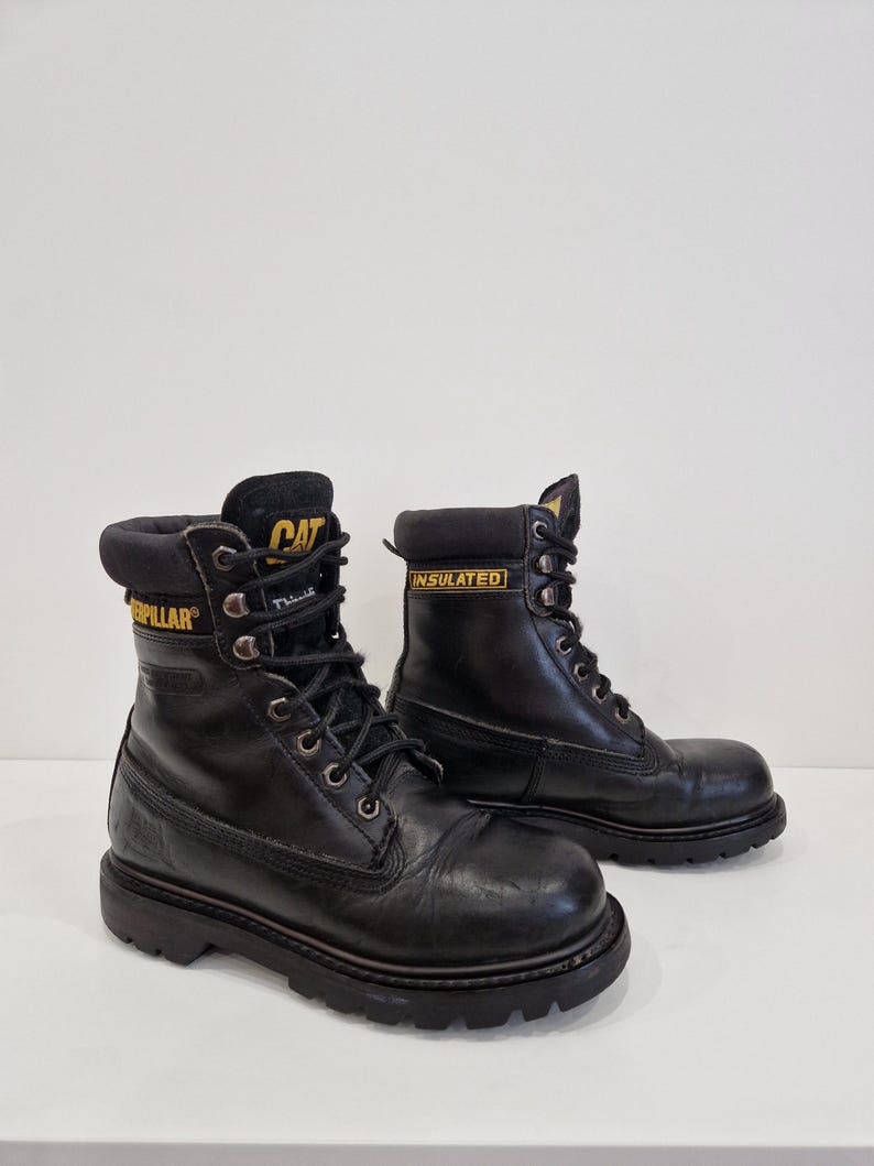 Puede incluir: Botas de trabajo de cuero negro con letras amarillas "CAT" e "INSULATED". Las botas tienen un dise&ntilde;o con cordones, ojales met&aacute;licos y una suela gruesa. Botas dise&ntilde;adas para el trabajo y actividades al aire libre.