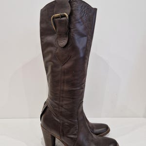 May include: Brown leather knee-high boots with a block heel and a decorative buckle. The boots have a rounded toe and a textured finish. The heel height is approximately 3 inches.