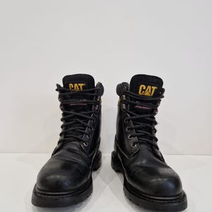 Puede incluir: Un par de botas de trabajo de cuero negro con la marca "CAT" en letras amarillas en la leng&uuml;eta. Las botas tienen cordones negros, ojales met&aacute;licos y una suela gruesa de goma negra. Las botas est&aacute;n dise&ntilde;adas para el trabajo y tienen una apariencia robusta.