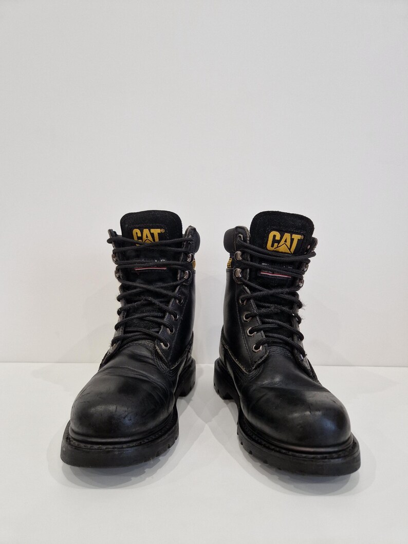 Puede incluir: Un par de botas de trabajo de cuero negro con la marca "CAT" en letras amarillas en la leng&uuml;eta. Las botas tienen cordones negros, ojales met&aacute;licos y una suela gruesa de goma negra. Las botas est&aacute;n dise&ntilde;adas para el trabajo y tienen una apariencia robusta.