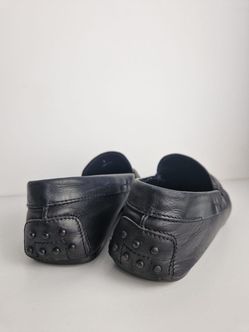 May include: Close-up of two black leather loafers. The shoes have a classic design with a rounded toe and a rubber sole with small, round studs. The leather appears to be smooth and slightly worn, giving the shoes a vintage look.