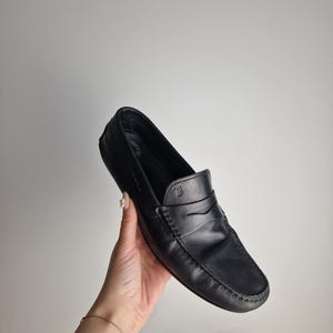 May include: Black leather loafer with a penny strap design. The shoe features a rounded toe and visible stitching details. The loafer is held by a hand against a plain white background. The shoe is a classic style.