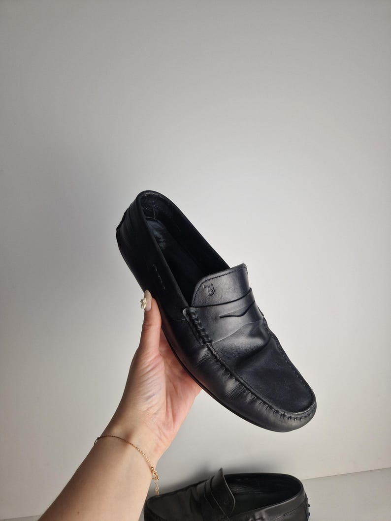 May include: Black leather loafer with a penny strap design. The shoe features a rounded toe and visible stitching details. The loafer is held by a hand against a plain white background. The shoe is a classic style.