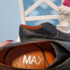 Puede incluir: Zapatos de vestir de cuero negro con detalles plateados. El interior es naranja con la palabra "MAX" en plata. Los zapatos tienen un dise&ntilde;o cl&aacute;sico con cordones, sobre fondo azul claro.