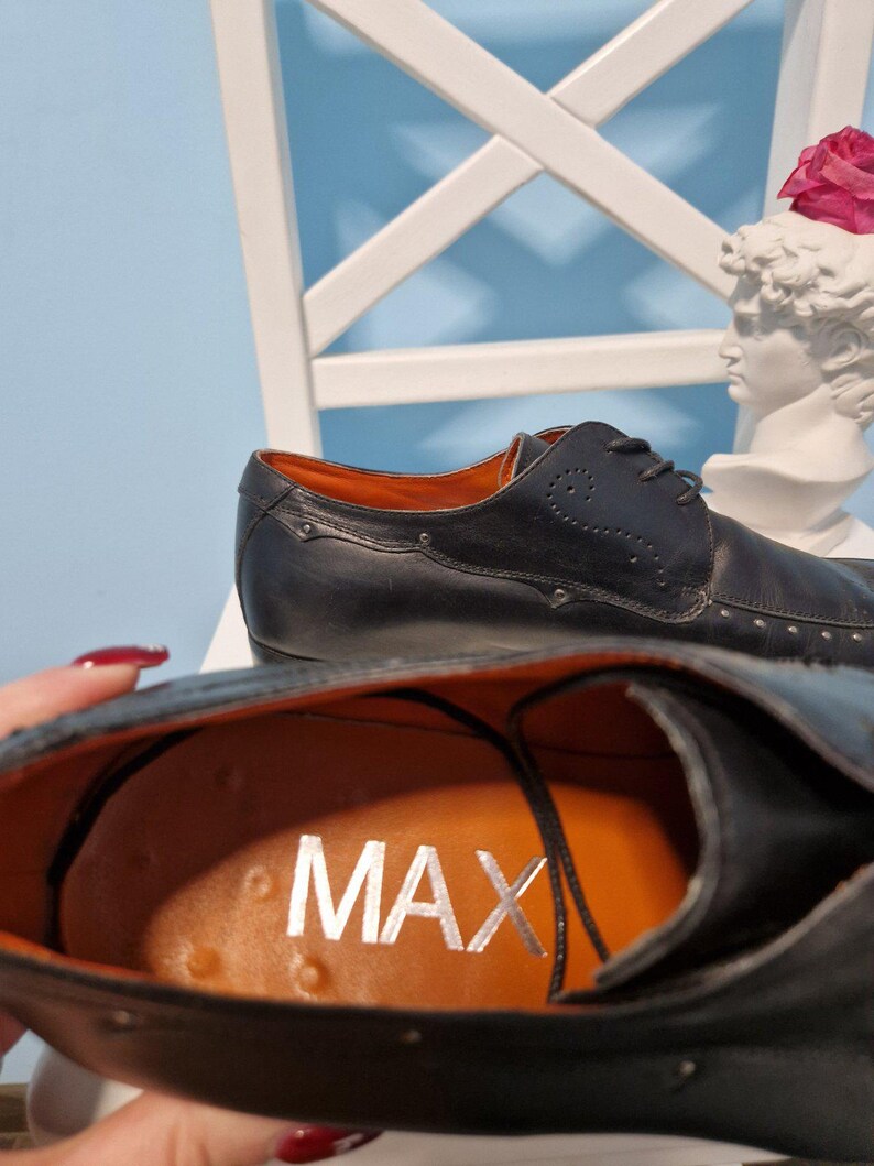 Puede incluir: Zapatos de vestir de cuero negro con detalles plateados. El interior es naranja con la palabra "MAX" en plata. Los zapatos tienen un dise&ntilde;o cl&aacute;sico con cordones, sobre fondo azul claro.
