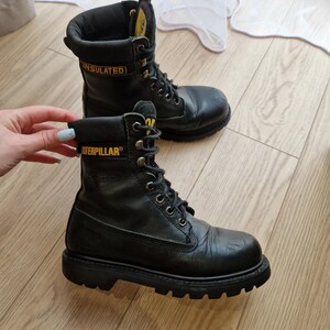 Puede incluir: Par de botas de trabajo Caterpillar negras con letras amarillas. Las botas son de cuero, con cordones y tienen una suela gruesa. La palabra "INSULATED" es visible en el lateral de una bota.