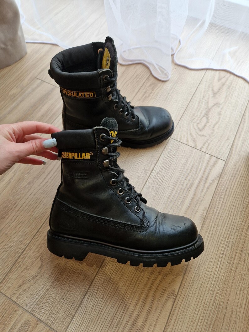 Puede incluir: Par de botas de trabajo Caterpillar negras con letras amarillas. Las botas son de cuero, con cordones y tienen una suela gruesa. La palabra "INSULATED" es visible en el lateral de una bota.