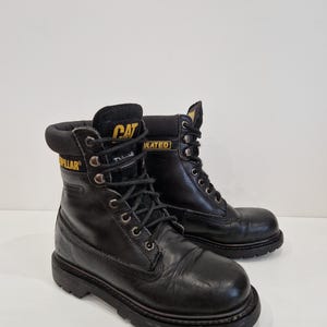 Puede incluir: Par de botas de trabajo Caterpillar negras. Las botas tienen un dise&ntilde;o con cordones y ojales met&aacute;licos, un cuello acolchado y una suela gruesa y resistente. El logotipo de Caterpillar es visible en amarillo en la leng&uuml;eta y el lateral de las botas.