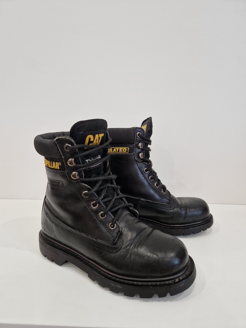 Puede incluir: Par de botas de trabajo Caterpillar negras. Las botas tienen un dise&ntilde;o con cordones y ojales met&aacute;licos, un cuello acolchado y una suela gruesa y resistente. El logotipo de Caterpillar es visible en amarillo en la leng&uuml;eta y el lateral de las botas.