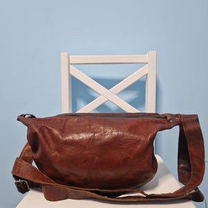 May include: A brown leather fanny pack with an adjustable strap. The bag is sitting on a white chair against a light blue wall. The leather has a textured appearance.