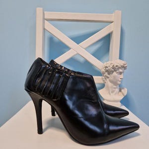 May include: A pair of black leather ankle boots with pointed toes and stiletto heels. The boots have a sleek, polished appearance and are set against a white chair and a light blue background. The heels are approximately 4 inches tall.