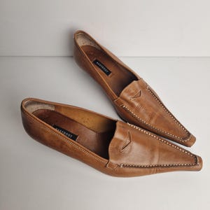 May include: A pair of tan leather loafers with a pointed toe and a low heel. The shoes feature a classic penny loafer strap across the vamp, visible white contrast stitching along the edges, and a black label reading NAVYBOOT inside the heel.