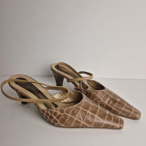 May include: A pair of beige slingback pumps with a textured crocodile-embossed pattern, featuring a pointed toe, a thin ankle strap, and a mid-height block heel. The interior insole is gold-toned and displays the brand name Peter Kaiser.
