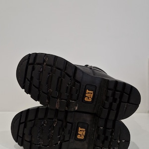 Puede incluir: Botas de trabajo negras con suela resistente, con el logotipo CAT en amarillo. Las botas est&aacute;n usadas, mostrando signos de uso. Las suelas tienen un dibujo profundo para agarre.