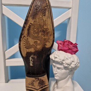 Puede incluir: Zapato de cuero negro con un dise&ntilde;o detallado de drag&oacute;n en la suela. Un busto blanco de David sostiene una rosa rosa. El zapato est&aacute; colocado en una silla blanca contra una pared azul claro.