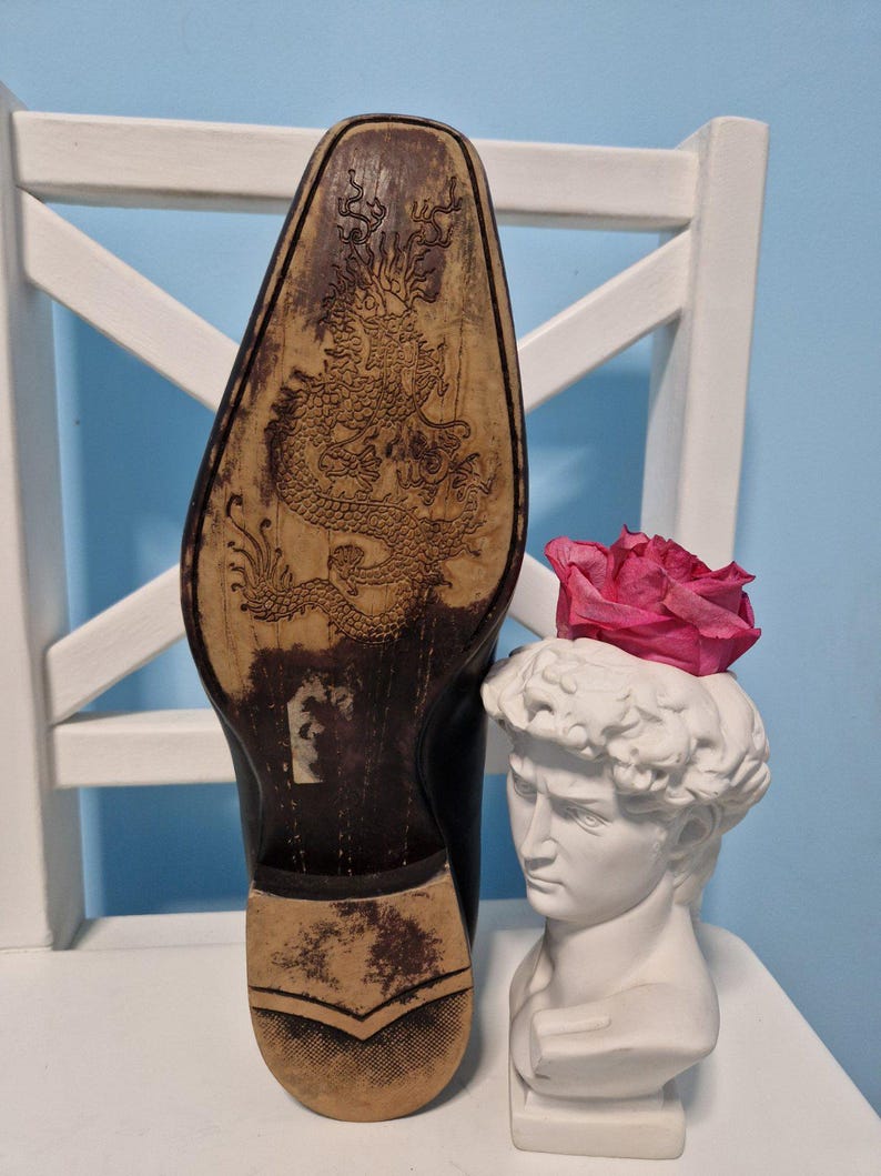 Puede incluir: Zapato de cuero negro con un dise&ntilde;o detallado de drag&oacute;n en la suela. Un busto blanco de David sostiene una rosa rosa. El zapato est&aacute; colocado en una silla blanca contra una pared azul claro.
