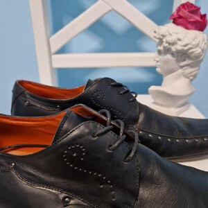 Puede incluir: Zapatos de vestir de cuero negro con costuras decorativas y tachuelas plateadas. Los zapatos tienen un dise&ntilde;o con cordones y un interior naranja. Un busto blanco con una flor rosa en el fondo.