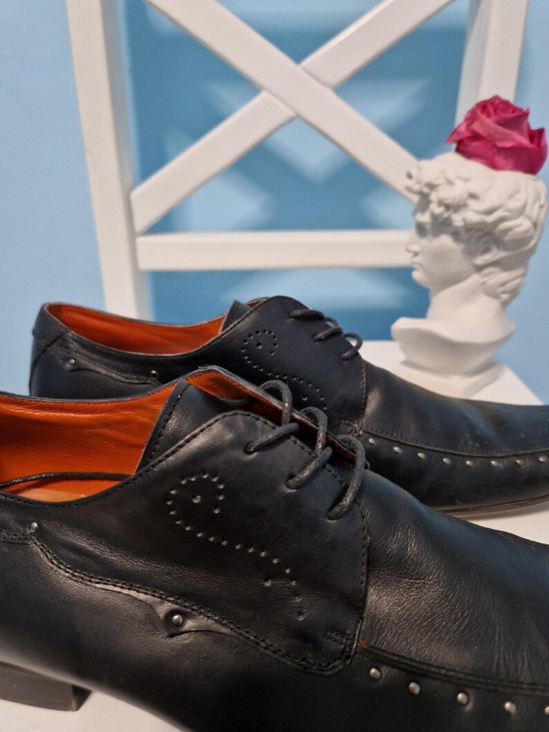 Puede incluir: Zapatos de vestir de cuero negro con costuras decorativas y tachuelas plateadas. Los zapatos tienen un dise&ntilde;o con cordones y un interior naranja. Un busto blanco con una flor rosa en el fondo.
