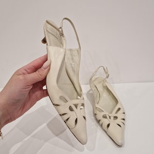 Puede incluir: Zapatos de tacón slingback de cuero blanco roto con puntera. Los zapatos tienen diseños calados decorativos en la pala y una fina correa en el tobillo. Los tacones miden unos 5 cm.