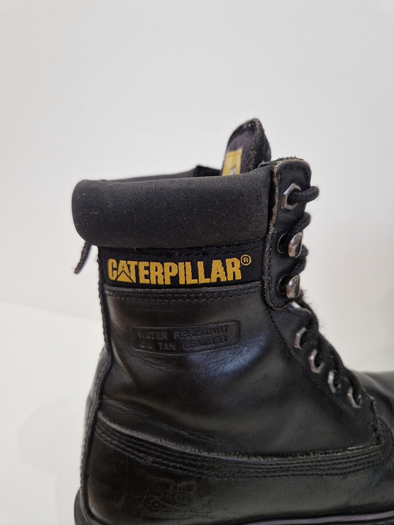 Puede incluir: Bota de trabajo de cuero negro con el logotipo "CATERPILLAR" en amarillo. La bota tiene ojales met&aacute;licos y un cuello acolchado. Las palabras "WATER RESISTANT" y "OIL TAN LEATHER" est&aacute;n grabadas en el lateral.