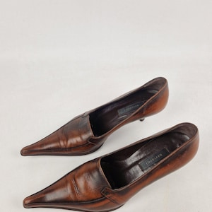 May include: A pair of brown leather pointed-toe pumps. The shoes have a low heel and a slip-on design. The interior of each shoe has a black label with white text. The shoes are set against a white background.