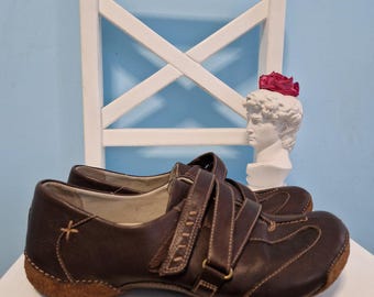 May include: A pair of brown leather shoes with two adjustable straps and a stitched design. The shoes have a light brown sole and a small embroidered detail on the side. The shoes are displayed on a white surface.