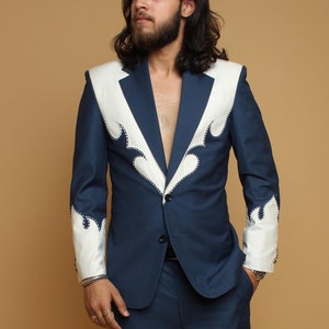 Peut inclure: Un homme portant un costume bleu foncé avec des bordures blanches sur les revers et les poignets. Le costume a une fermeture à bouton unique et l'homme porte des chaussures noires.