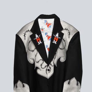 May include: Black blazer with silver accents and barbed wire embroidery. The blazer features a white satin lapel and cuffs with decorative stitching. The interior lining is white with a repeating graphic of a cowboy and the letters "R" and "H".