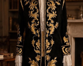 May include: A long black coat featuring intricate gold embroidery in a scrolling leaf pattern. The coat has a high collar, long sleeves with white lace cuffs, and is displayed on a mannequin. A white lace shirt is visible underneath the open front.