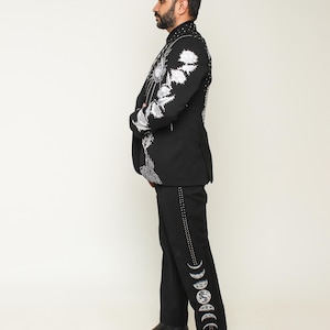 May include: Black suit with intricate white floral embroidery on the jacket and moon phase designs on the pants. The suit features a high collar and is paired with black leather boots. The overall style is formal and unique.