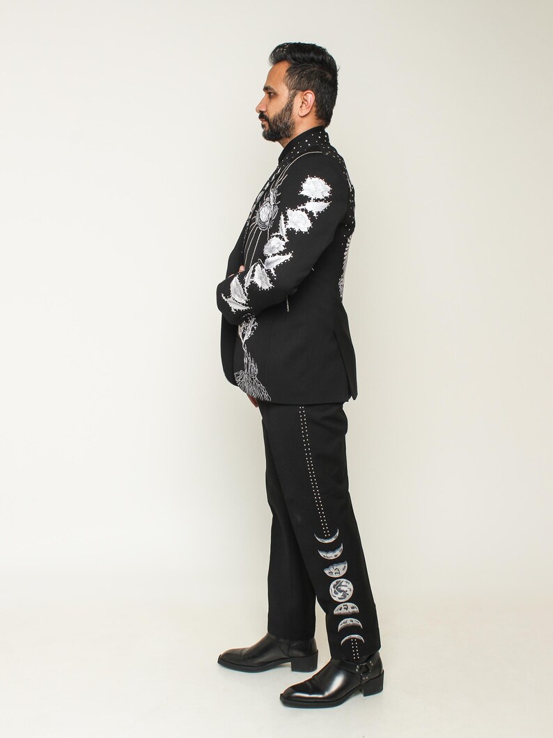May include: Black suit with intricate white floral embroidery on the jacket and moon phase designs on the pants. The suit features a high collar and is paired with black leather boots. The overall style is formal and unique.