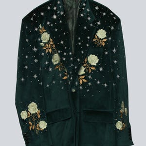Puede incluir: Una blazer de terciopelo verde oscuro con bordados de rosas blancas y hojas doradas. La blazer tiene un cierre de un solo bot&oacute;n, dos bolsillos delanteros y est&aacute; adornada con adornos en forma de estrella. Las mangas tienen detalles de botones.
