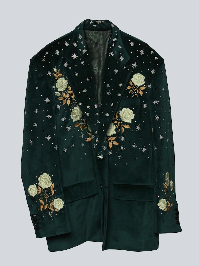 Puede incluir: Una blazer de terciopelo verde oscuro con bordados de rosas blancas y hojas doradas. La blazer tiene un cierre de un solo bot&oacute;n, dos bolsillos delanteros y est&aacute; adornada con adornos en forma de estrella. Las mangas tienen detalles de botones.