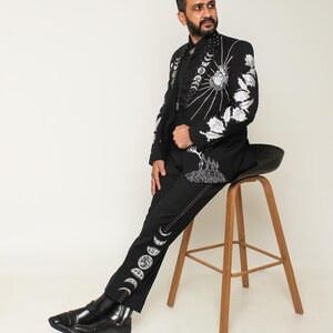 May include: Black suit with intricate white embroidery featuring celestial and floral designs. The jacket and pants are adorned with detailed patterns, including a sunburst, moon phases, and floral motifs. The suit is paired with black boots.