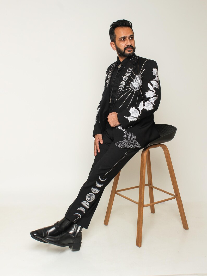 May include: Black suit with intricate white embroidery featuring celestial and floral designs. The jacket and pants are adorned with detailed patterns, including a sunburst, moon phases, and floral motifs. The suit is paired with black boots.
