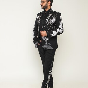 May include: Black suit with intricate white embroidery featuring celestial and floral designs. The jacket has a detailed pattern on the sleeves and lapels. The pants have a decorative pattern down the side. The suit is paired with a black shirt and black shoes.