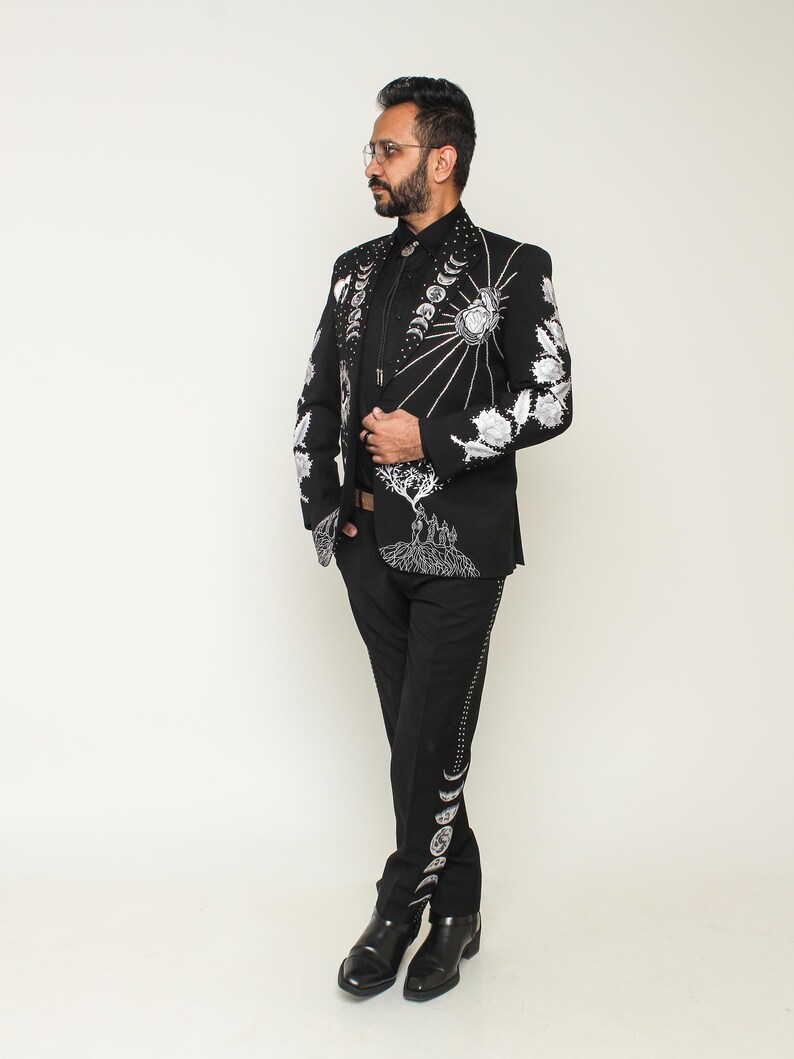 May include: Black suit with intricate white embroidery featuring celestial and floral designs. The jacket has a detailed pattern on the sleeves and lapels. The pants have a decorative pattern down the side. The suit is paired with a black shirt and black shoes.