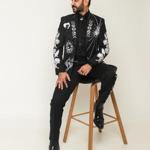 May include: Black suit jacket with intricate white celestial embroidery, including moons, suns, and stars. The jacket is paired with matching black pants and black boots. The person is sitting on a wooden stool.