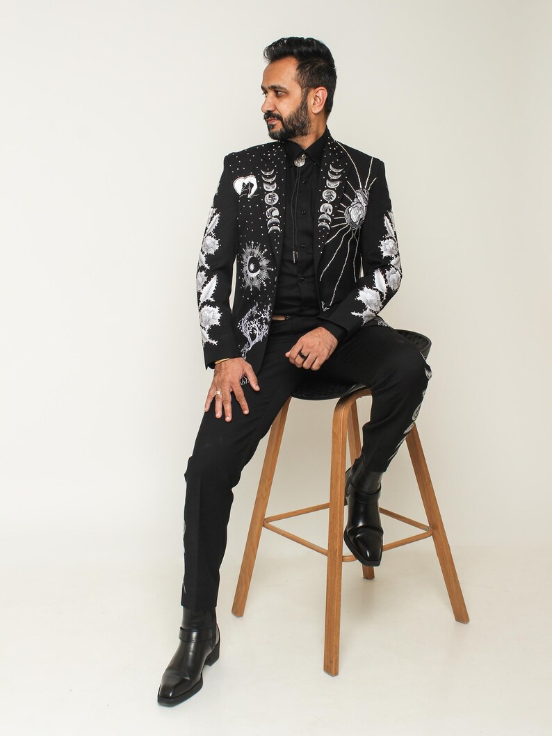May include: Black suit jacket with intricate white celestial embroidery, including moons, suns, and stars. The jacket is paired with matching black pants and black boots. The person is sitting on a wooden stool.